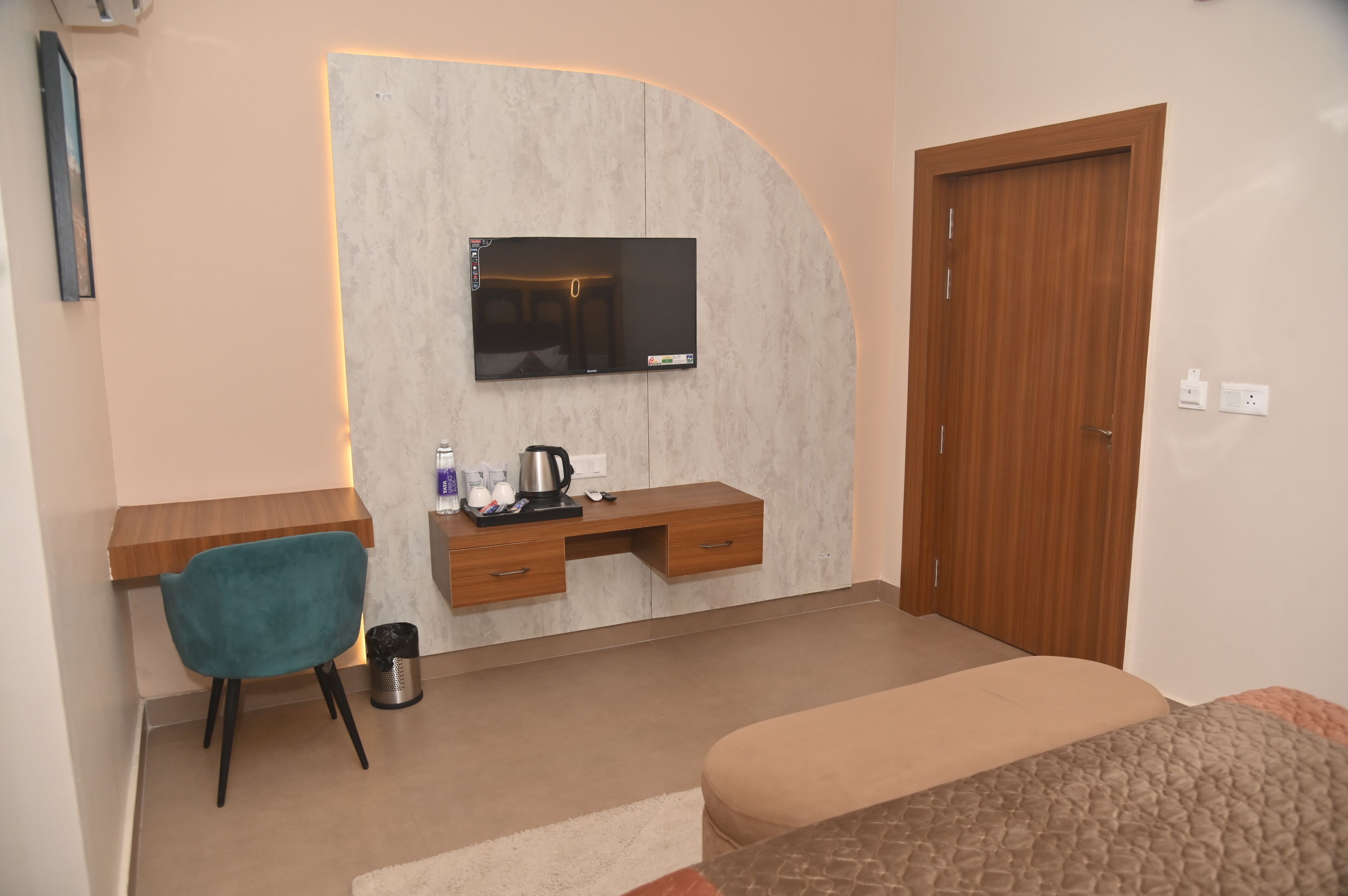 A modern bedroom with a TV mounted on a curved wall, a wooden desk, a teal chair, a bed, and a wooden door.