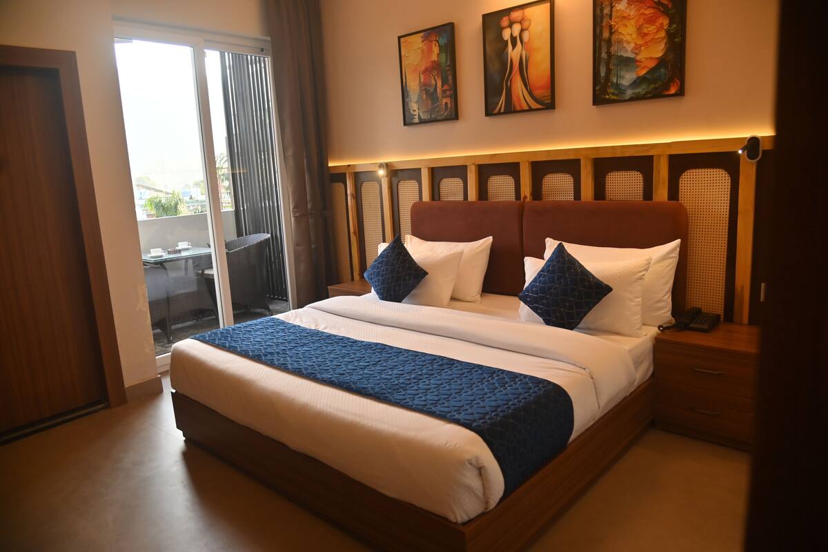 A hotel room with a large bed, a wooden headboard, a small table, and a chair. There are two paintings on the wall above the bed.