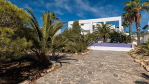 A modern building with a stone pathway leading to a pool area, surrounded by palm trees and other vegetation. - Villa 'Casa Arola' with Sea Views, Wi-Fi and Air Conditioning (Sant Josep de sa Talaia)