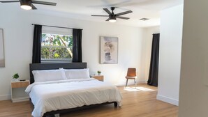 Ein modernes Schlafzimmer mit Bett, Deckenventilatoren, einem Fenster mit Vorhängen, einem Nachttisch mit Pflanze und Wandkunst.