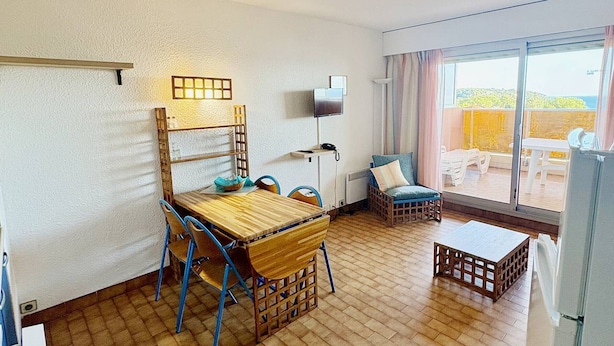 Une pièce comprenant une table à manger en bois, des chaises bleues, un fauteuil en osier, une petite table en bois et une vue sur l’extérieur grâce à une grande fenêtre.