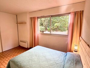 Ein kleines, schlichtes Zimmer mit einem Bett, einem Fenster mit Vorhängen, einem Heizkörper und einer Lampe.