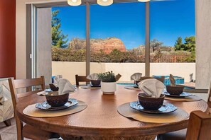 A dining table with four bowls and napkins, a potted plant, and a view of a red rock formation through large windows. - Peaceful Sedona Retreat, Views, Pool, Hot Tub! (Sedona)