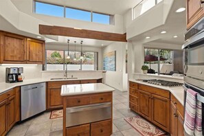 A modern kitchen with wooden cabinets, a stainless steel dishwasher, a large island with drawers, a gas stove, and a view of the outdoors through large windows. - Peaceful Sedona Retreat, Views, Pool, Hot Tub! (Sedona)