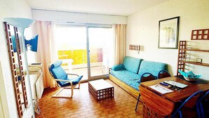 Ein Wohnzimmer mit Sofa, Sessel, Tisch und einem Balkon mit gelanderfarbener Umrandung.