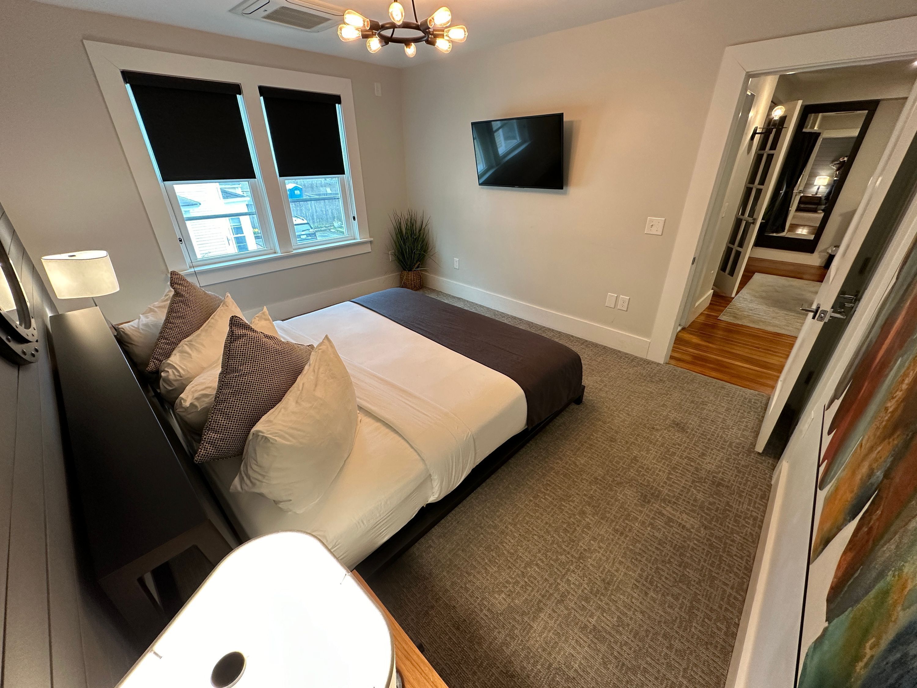 Een moderne slaapkamer met een bed, een flatscreen-tv, een raam met jaloezieën, een plafondlamp en een gang naar een andere kamer.