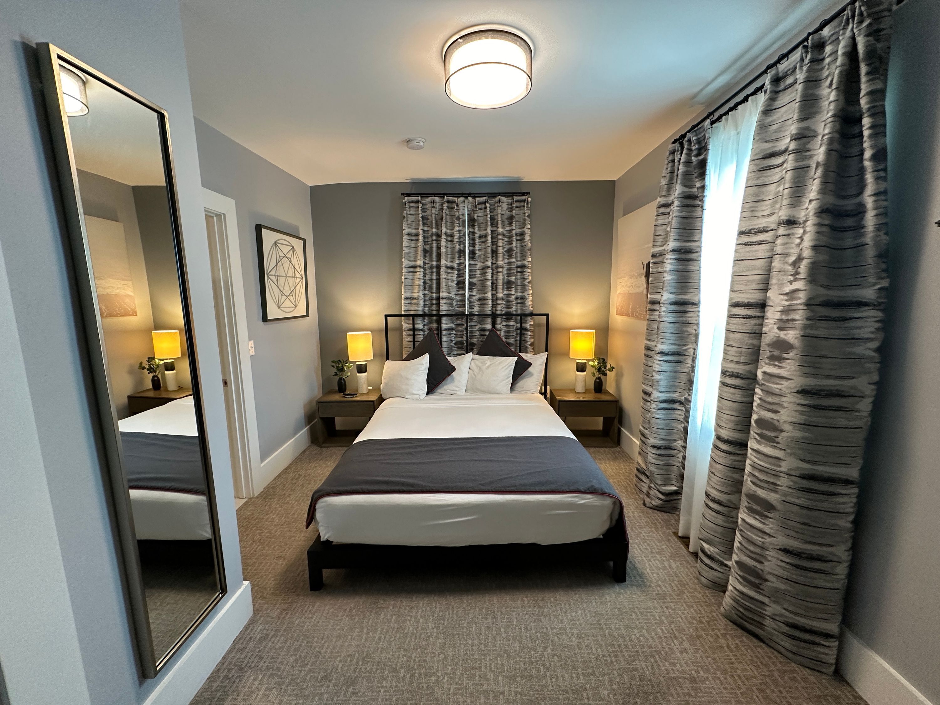 A hotel room with a large bed, two bedside tables with lamps, a ceiling light, a large mirror, and a window with curtains.