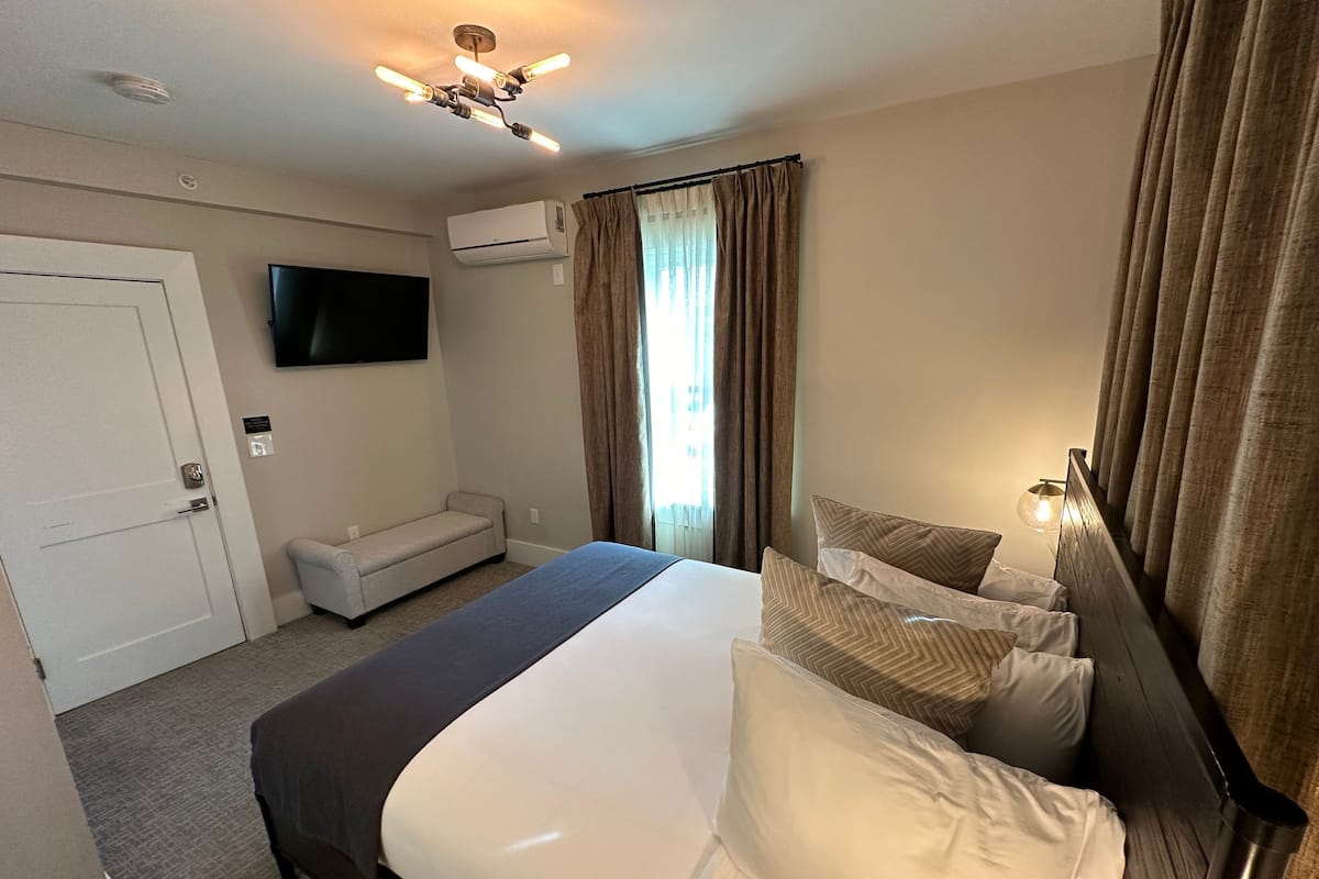 Una habitación de hotel con cama, televisor, aire acondicionado, una banca y una ventana con cortinas.