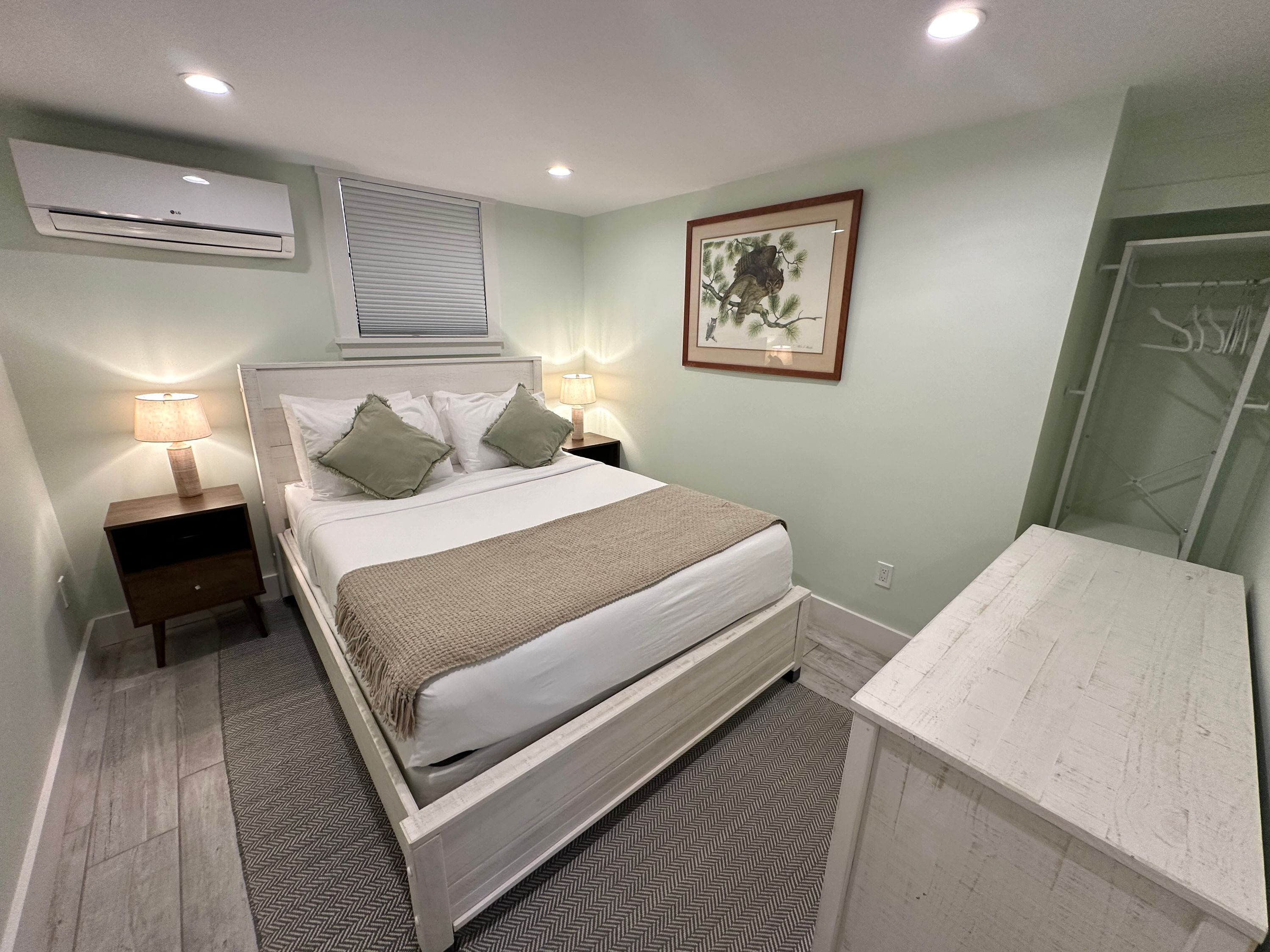 Een slaapkamer met een bed, nachtlampjes, een nachtkastje, een airconditioner, een kunstwerk in een lijst en een garderobe.