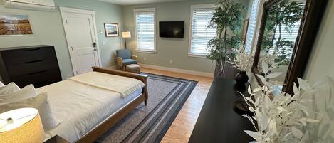 Um quarto com cama, mesa de cabeceira, luminária, cômoda, cadeira, televisão, janelas e ventilador de teto.