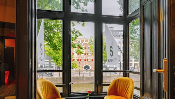 A room with two yellow chairs, a small table, and a view of a building with a sign that reads "Superhotel." - Supper boutique suites (Amsterdam)
