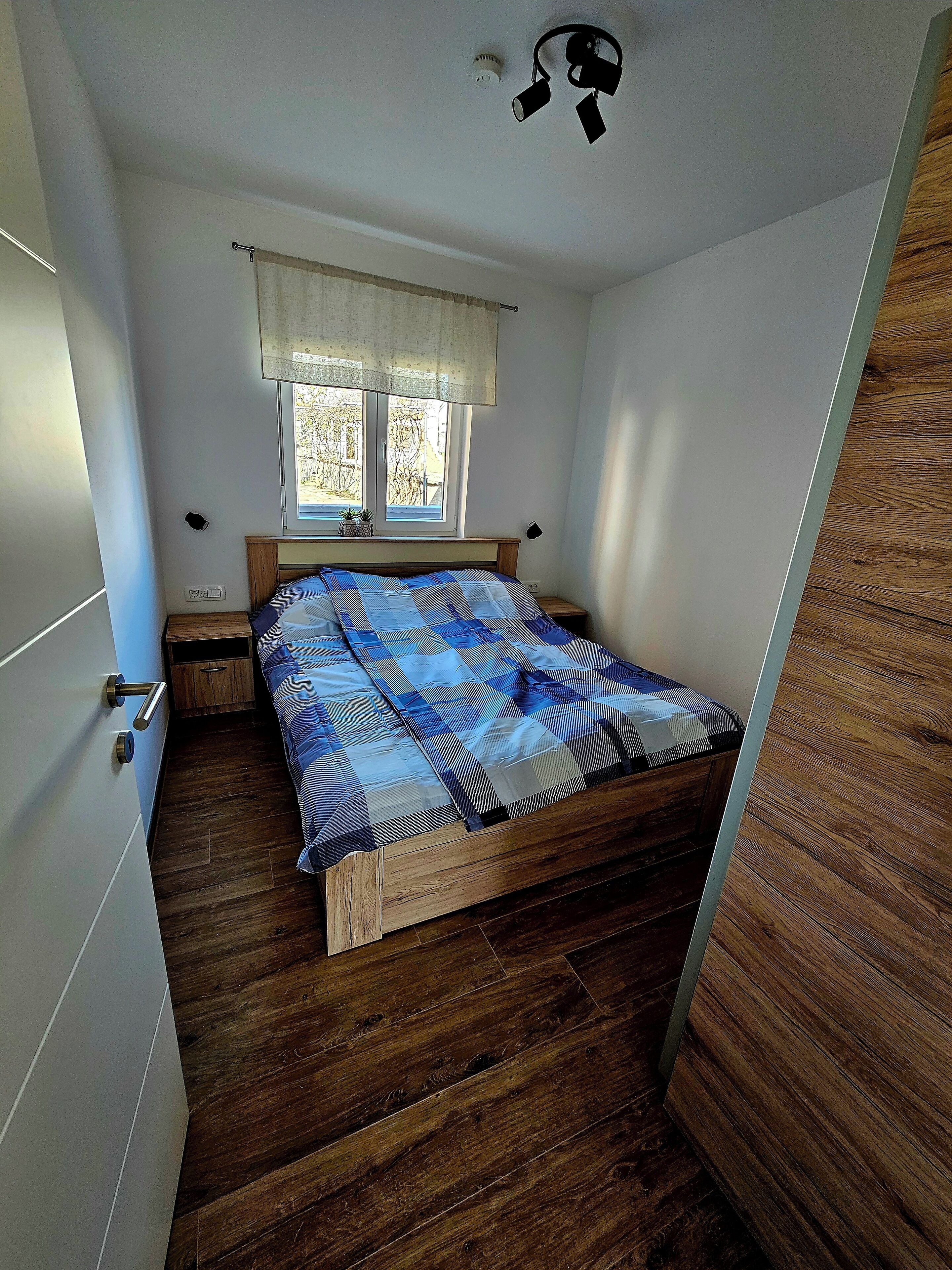 A bedroom with a wooden bed, a window with a curtain, a bedside table, and a nightstand.