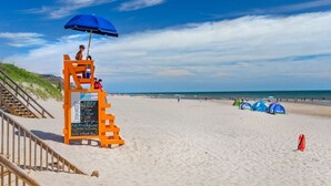 Ein Rettungsschwimmturm mit blauem Sonnenschirm, einer Holztreppe und einem sandigen Strand mit Zelten.