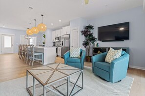 A modern living room with a blue sofa set, a dining area with chairs, a kitchen with white cabinets, and a flat-screen TV mounted on the wall. - Ebb Tide 3 Bedroom at Cambridge Cove with Private Hot Tub! (Kill Devil Hills)