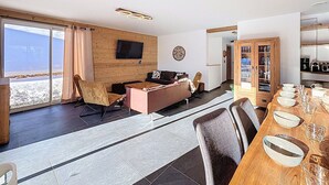 Un salon moderne avec un mur en bois, un téléviseur à écran plat, un coin repas comprenant une table et des chaises en bois, et une vue sur l’extérieur.