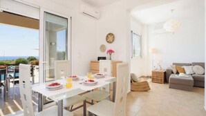 A modern living room with a sofa, a dining table set with plates and glasses, and a view of the sea through large windows. - Villa Nicole Gennadi (2W76+JJ South Rhodes, Greece)