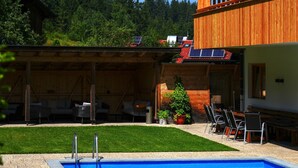 A pool area with a covered seating area, a wooden building, and a view of a forested hillside. - Aparthotel Iris by Interhome (Auffach)