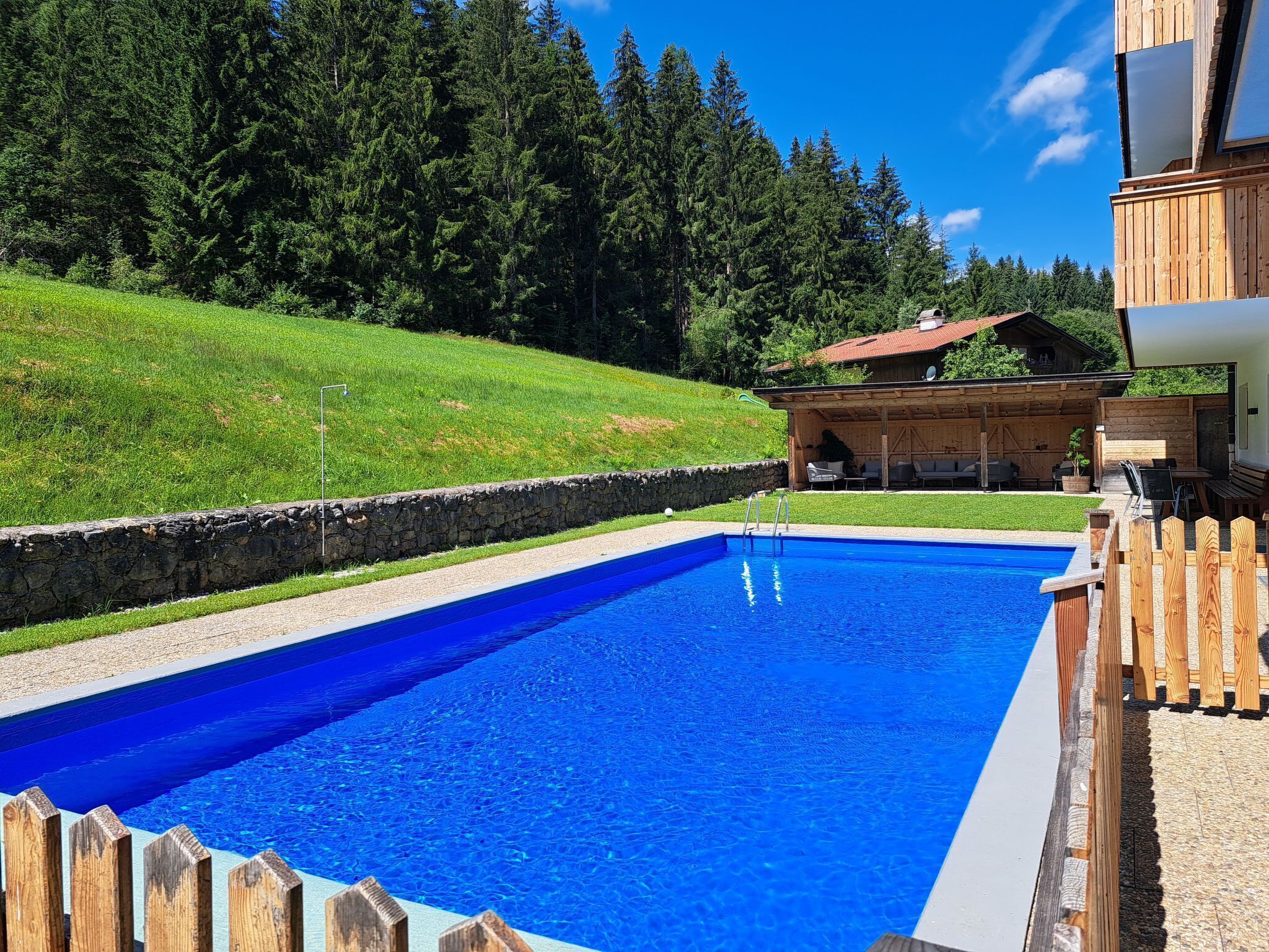 A swimming pool with a stone wall and wooden deck, surrounded by a grassy area and a wooden building with a red roof.