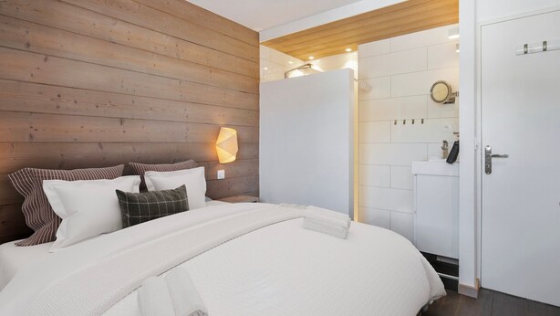 Een slaapkamer met een houten hoofdbord, een bed met wit beddengoed, een bedlampje, en een badkamer zichtbaar door een open deur.