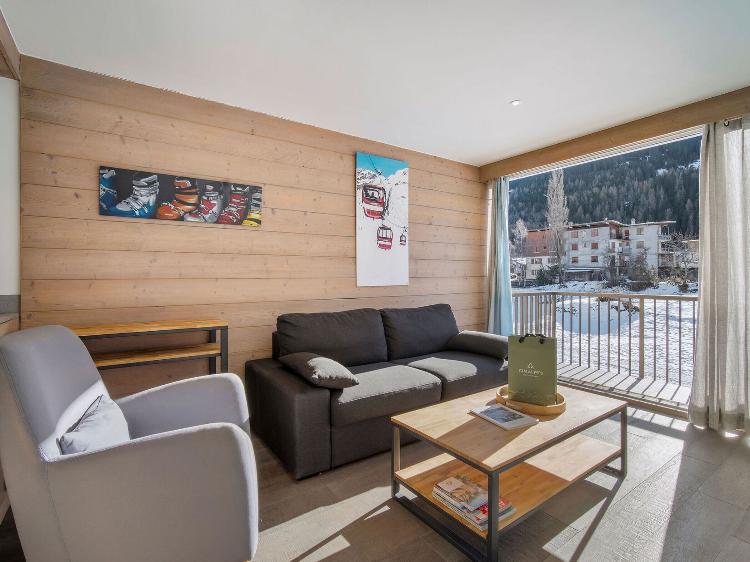 A modern living room with a sofa, armchair, and coffee table. There are two paintings on the wall and a view of a snowy landscape through a large window.