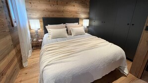 Ein Schlafzimmer mit Holzbalkendecke, einem Bett mit weißer Bettwäsche, einem Nachttisch mit Lampe und einem dunklen Kleiderschrank.