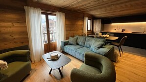 Ein gemütliches Blockhaus-Zimmer mit Holzbalkendecke, einer Couch, zwei Sesseln, einem kleinen Tisch und einer Küchenzeile mit Esstisch und Stühlen.