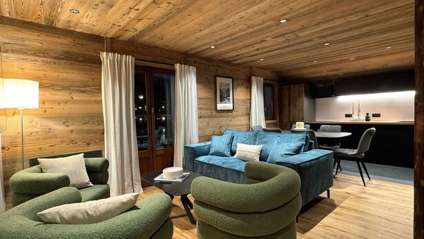 Ein gemütliches Blockhaus-Interieur mit einer Couch, Sesseln, einem Esstisch und einer Küchenzeile.