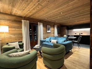 A cozy log cabin interior with a sofa, armchairs, a dining table, and a kitchen area. - Beautiful T4 apartment in the village center, close to slopes, parking (La Clusaz)