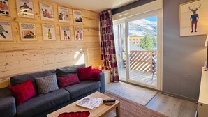 Ein gemütliches Wohnzimmer mit einer grauen Couch, roten und grauen Kissen, einem Holztisch mit einer Zeitschrift und roten Schalen, sowie einem großen Fenster mit Blick auf Berge und eine Holzterrasse davor.
