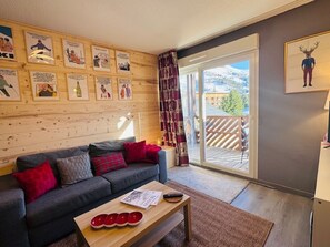 A cozy living room with a grey sofa, red and grey cushions, a wooden coffee table with a magazine and red bowls, and a large window with a view of mountains and a wooden deck outside. - Renovated apartment in central Les 2 Alpes, near slopes (Les Deux Alpes)