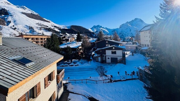Ein schneebedecktes Dorf mit traditioneller Bebauung, blauem Himmel und Bergen im Hintergrund.