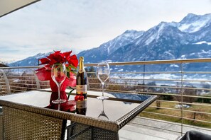 Ein Tisch mit einer Weinflasche, einem Glas und einem Strauß roter Blumen, vor einer Bergkulisse.