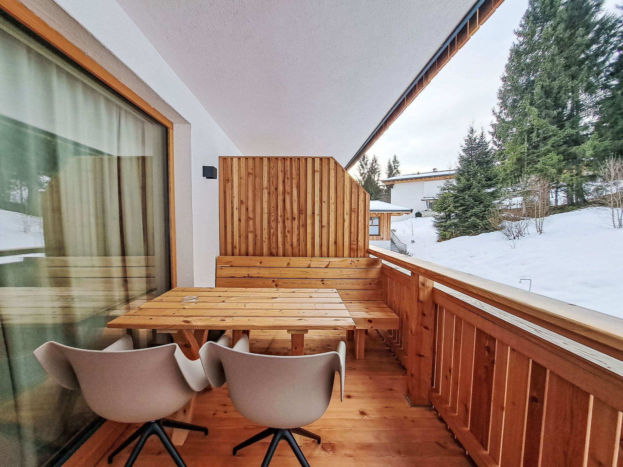 Eine Holzterrasse mit Tisch und Stühlen, mit Blick auf eine verschneite Landschaft und ein Gebäude mit Holzfassade.