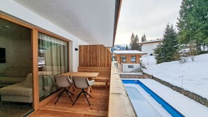 A wooden deck with a table and chairs, a glass door leading to a room, and a view of a snowy landscape with a building in the background. - Aparthotel Iris by Interhome (Auffach)