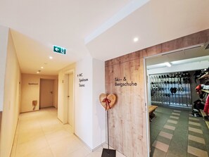 Un couloir d’hôtel avec des panneaux indiquant « Spielraum » et « Sauna », ainsi qu’un espace « Ski & Bergschuhe » où sont entreposés des chaussures et des bottes de ski.