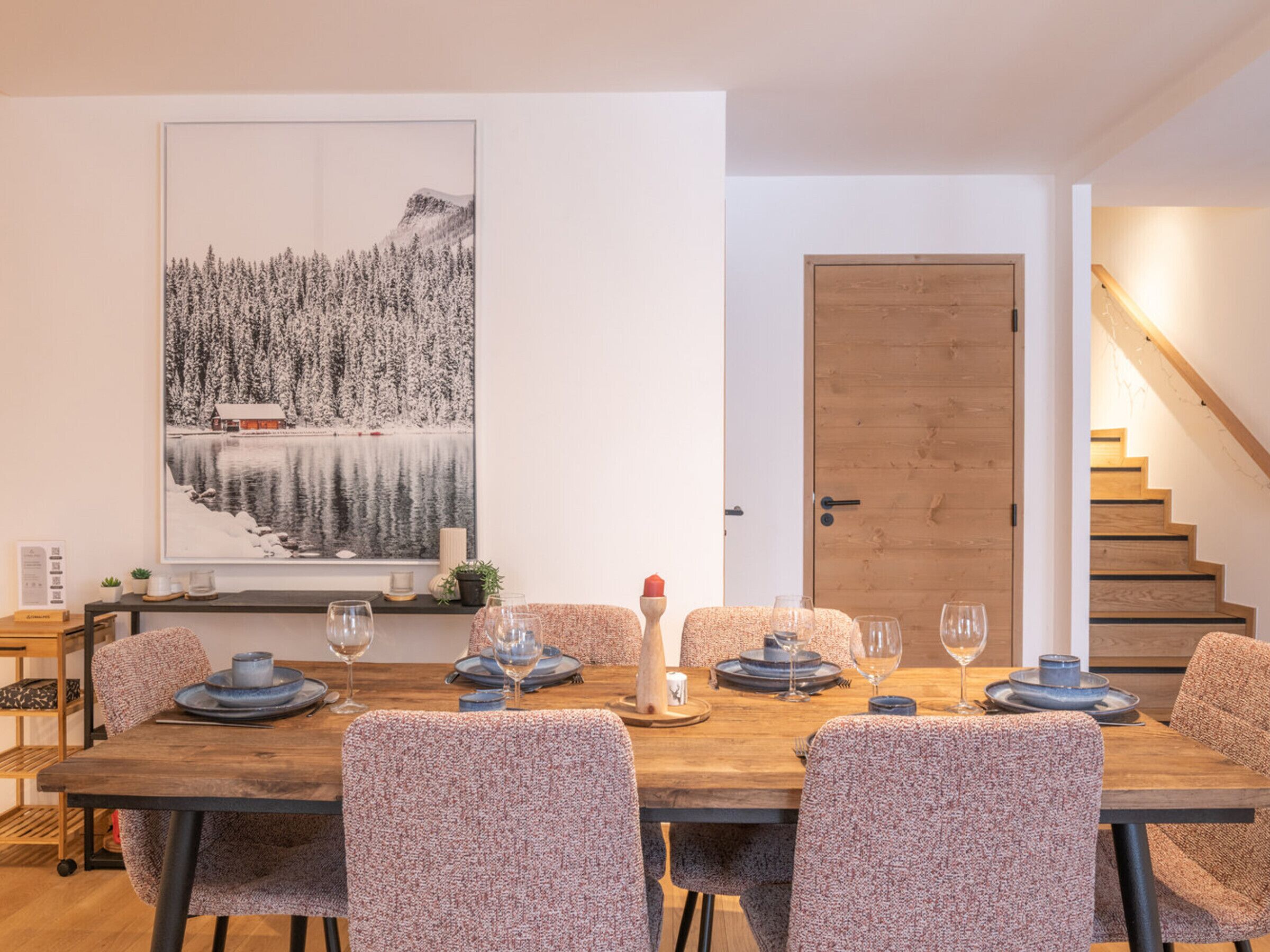 En matplats med ett träbord dukat för sex personer, omgiven av stolar med mönster. Ett stort, inramat fotografi av ett snöigt landskap hänger på väggen.