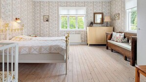 A bedroom with a wooden floor, a bed with a white headboard, a wooden bench with cushions, a dresser, and a window with a view of greenery.