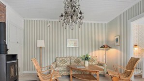 A cozy living room with a fireplace, striped wallpaper, a chandelier, a sofa, two wicker chairs, and a coffee table.