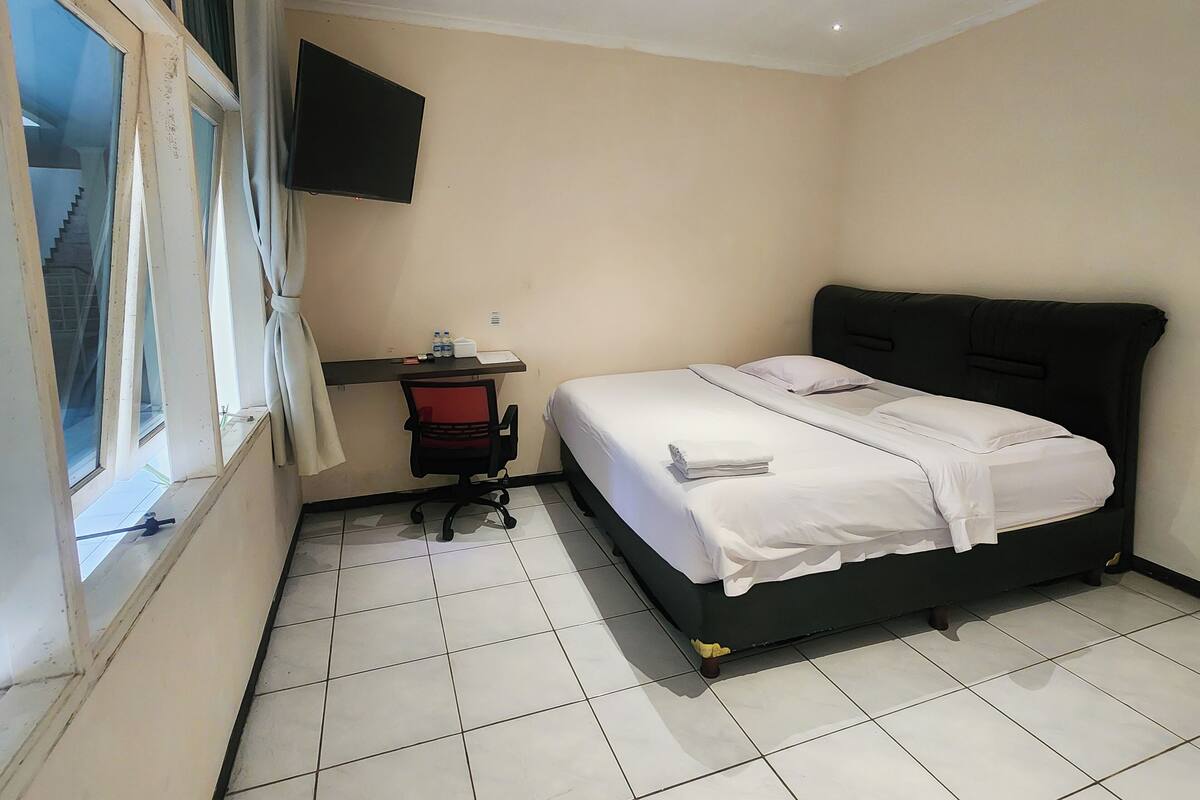 A hotel room with a bed, a desk, a chair, a TV mounted on the wall, and a window with curtains.