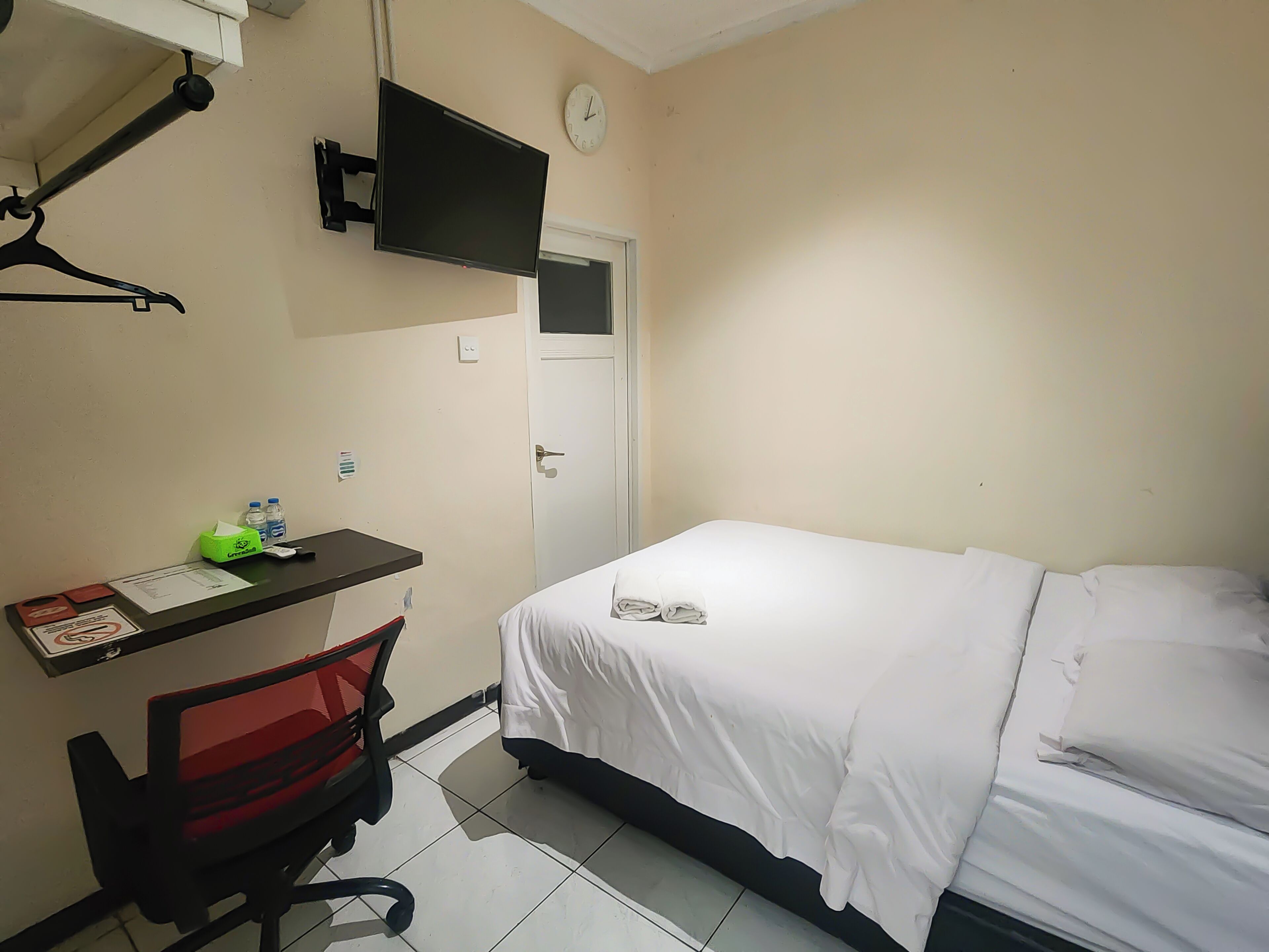 A hotel room with a bed, a desk with a chair, a TV mounted on the wall, and a door.