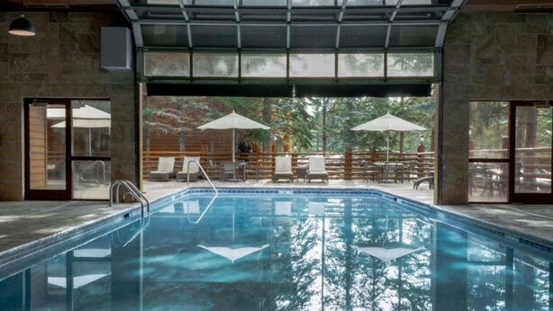 Piscina con agua cristalina, rodeada de sillones de descanso y sombrillas, con vista a árboles y una terraza de madera.