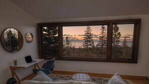 Ett rum med ett stort fönster, en bärbar dator på ett skrivbord, en soffa och ett litet bord med en lampa.