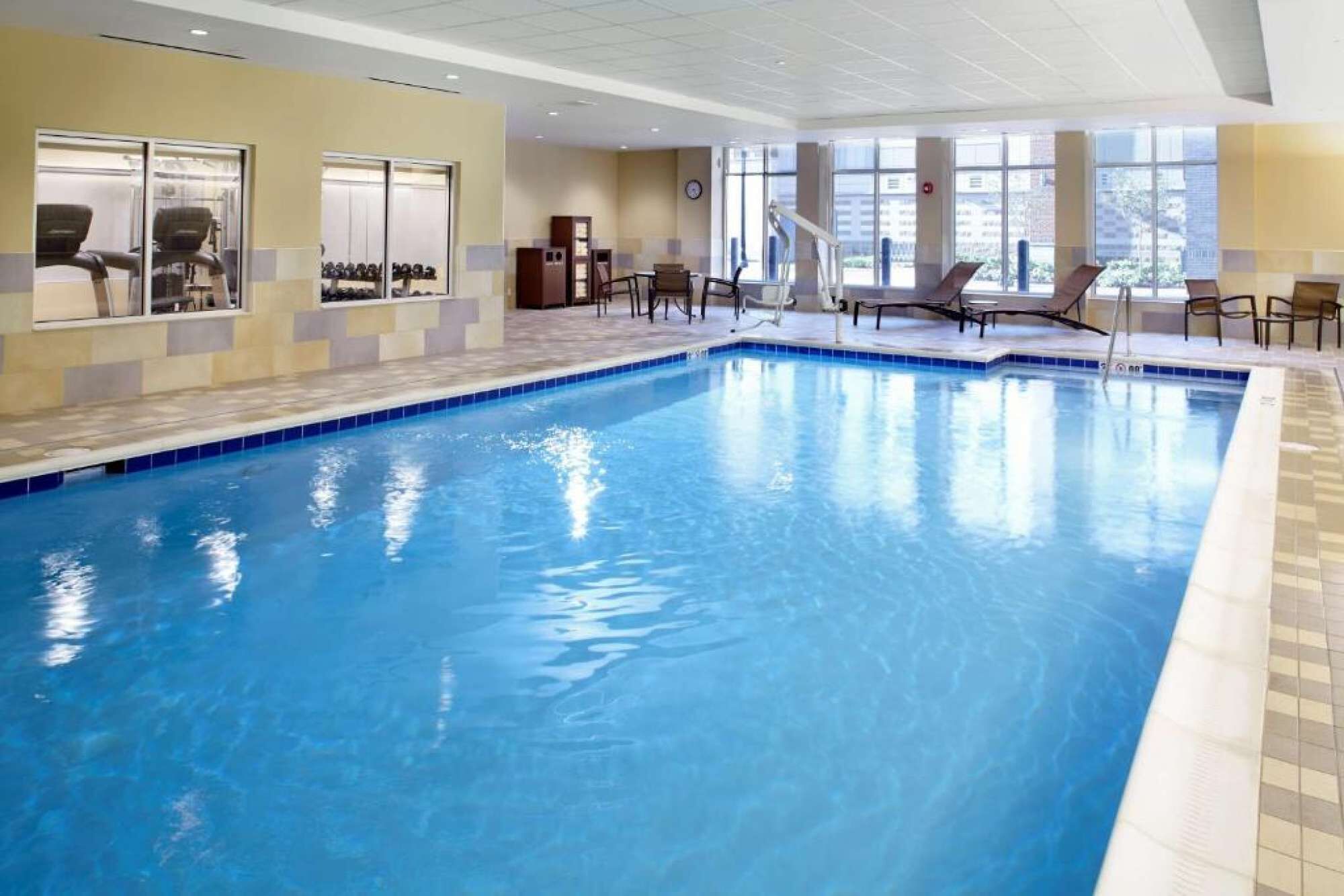An indoor swimming pool with a tiled edge, lounge chairs, and a view of the outside through large windows.