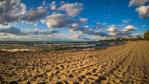 Une vaste plage de sable fin, parsemée d’empreintes menant à l’eau, un ciel dégagé avec quelques nuages, et une côte lointaine où l’on aperçoit des bâtiments.