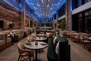 Een ruime, overdekte eetruimte met verschillende zithoeken, waaronder ronde tafels en banken, en een hoge plafond met decoratieve verlichting.