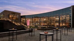 A modern building with a glass facade, outdoor seating area with round tables, and a staircase leading to an upper level. - By Gas South Convention Center + Rooftop Bar. Pool (Duluth)