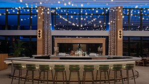 A bar area with a central bar counter, multiple stools, and a backdrop of hanging light bulbs. - By Gas South Convention Center + Rooftop Bar. Pool (Duluth)