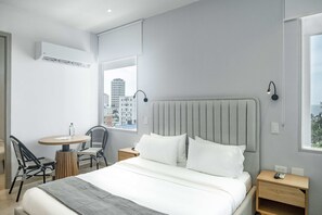Ein modernes Hotelzimmer mit Bett, Nachttischen, einem Sessel, einem kleinen Tisch und Blick auf die Stadt durch das Fenster.