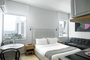 Ett modernt hotellrum med en säng, sängbord, en soffa, en matplats och utsikt över stadsbilden.