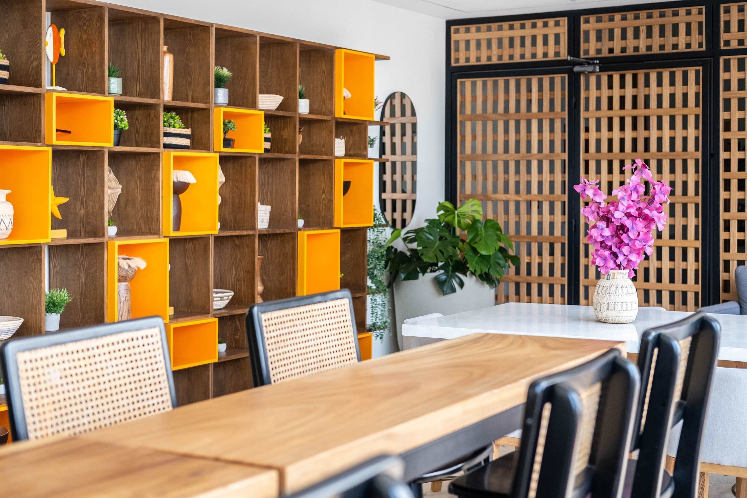 Ein Essbereich mit einem Holztisch und Stühlen, einem Bücherregal mit orangefarbenen und hölzernen Ablagen und einer Kübelpflanze mit rosa Blüten.